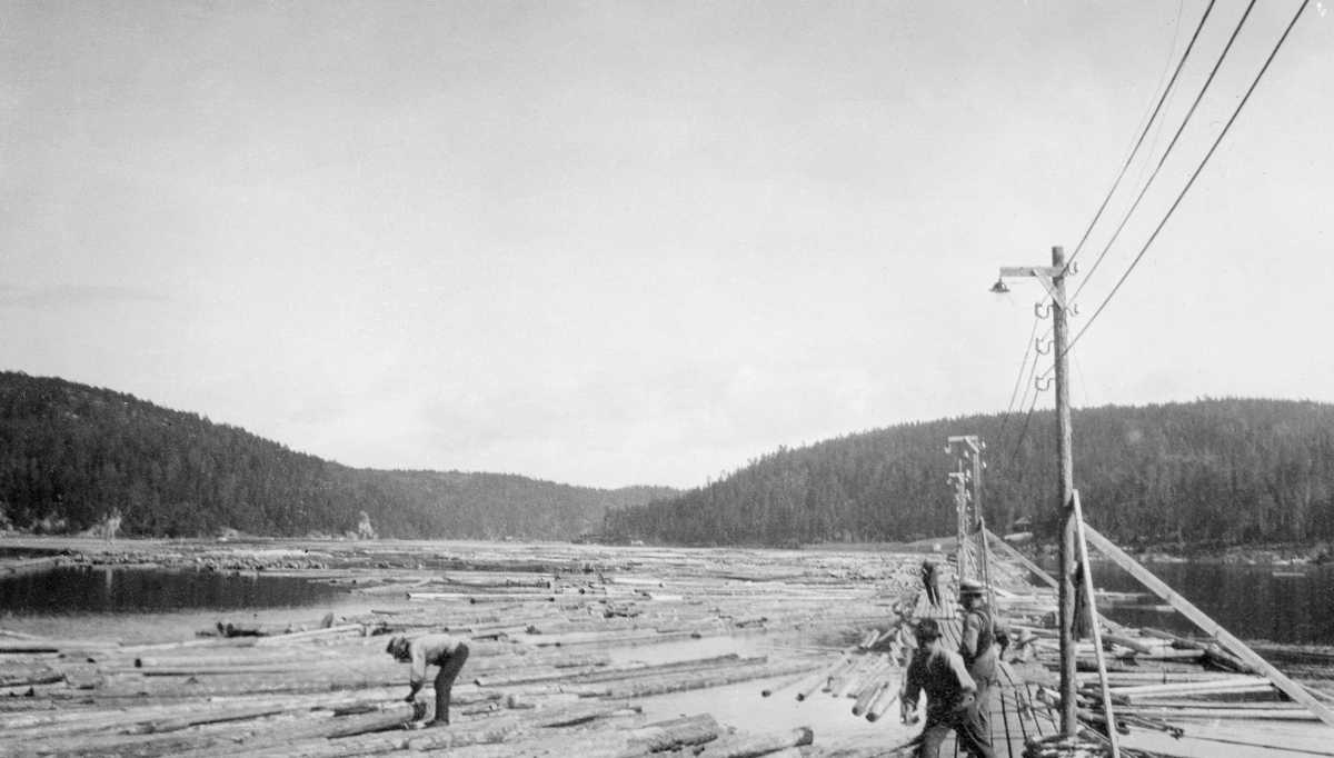 Fra påstiklingslensa ved kraftverksdammen i Solbergfoss i Askim i Østfold (nedre Glomma).  Fotografiet er tatt fra den ene lensearmen, den østre, som skulle hindre at tømmeret drev inn i Lierevja (mot sørøst).  Vi ser fløtere som arbeider fra flåtegangen, og vi ser også en mann som har vågd seg ut på det flytende løstømmeret, der han står bøyd over en arbeidsoppgave.  Bak på flåtegangene er det reist lyktestolper med elektriske lamper.  De gjorde det mulig å arbeide lange dager med påstikking også seinhøstes, når mørket senket seg tidlig på ettermiddagen. 