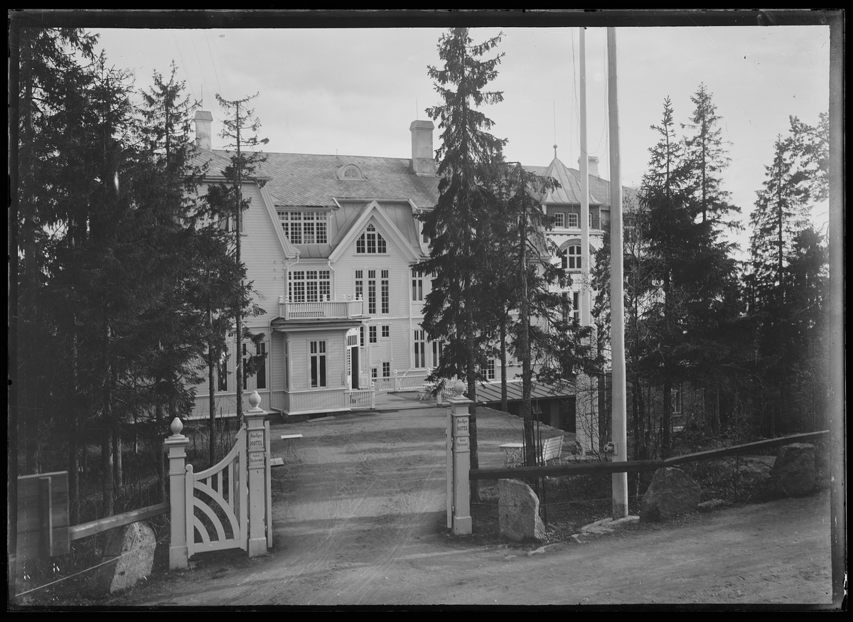 Fasaden til Anna Kruses Hotell, sett fra porten til hotellet. Det er grantrær på hver side av porten og to flaggstenger til høyre.