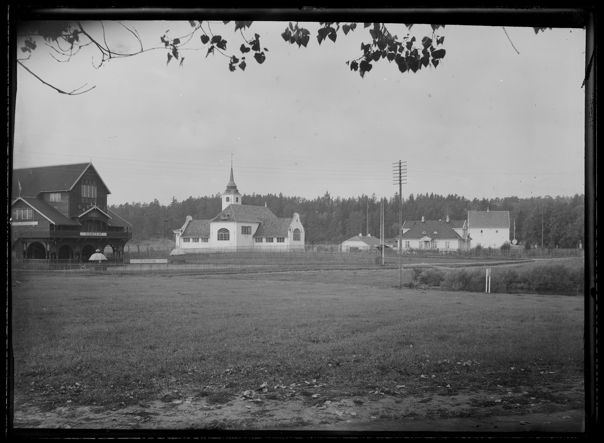 Utsikt fra et jorde til en hvit kirke og en konsertbygning med utendørs restaurant til venstre ved siden av. Trebygninger og skog sees i bakgrunnen.