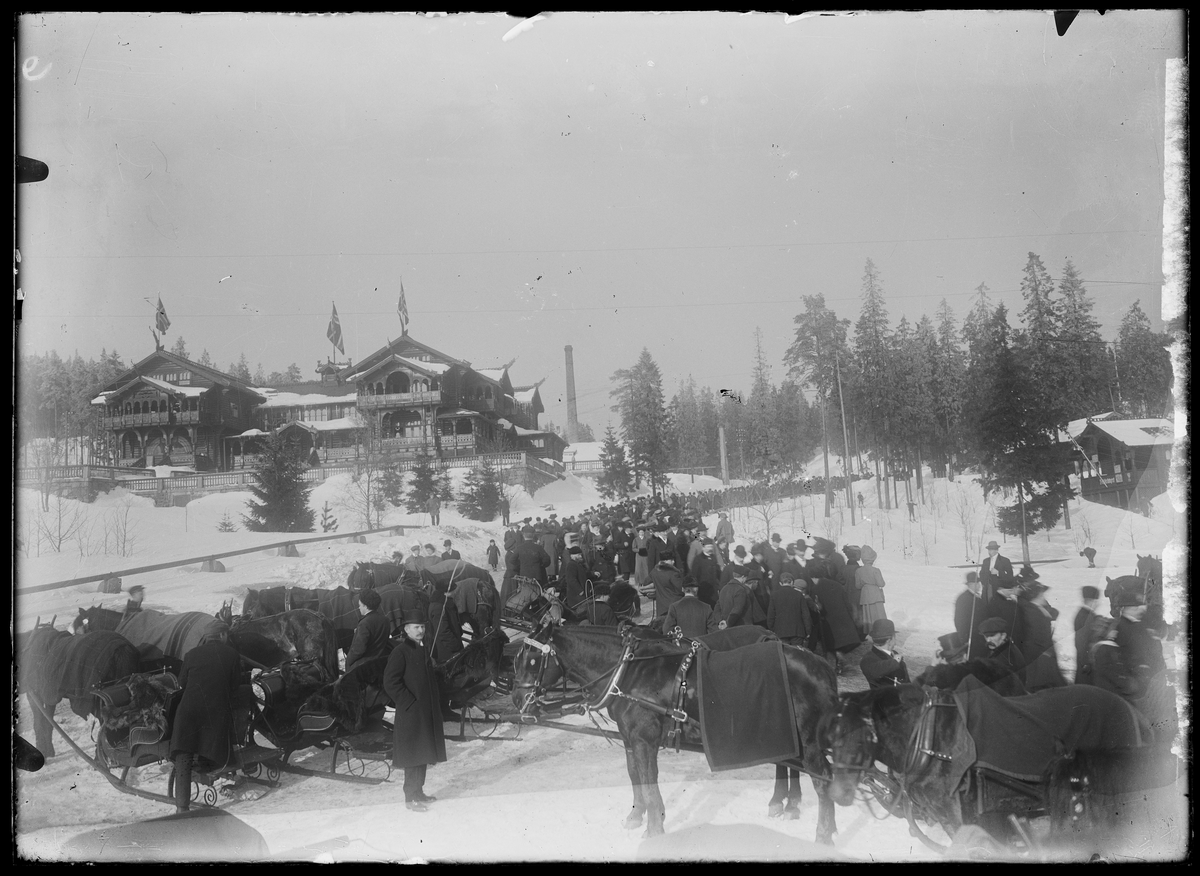 Utsikt til et hotell på en høyde i et skoglandskap. I forgrunnen er det en vrimmel av folk, hester og sleder.