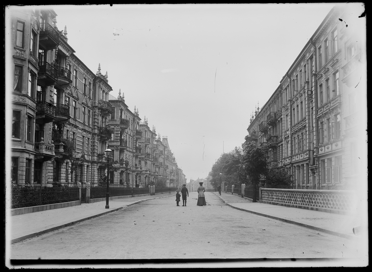 En gate går gjennom et bylandskap med bygårder og fortau på hver side. En kvinne og to barn står midt i gaten.