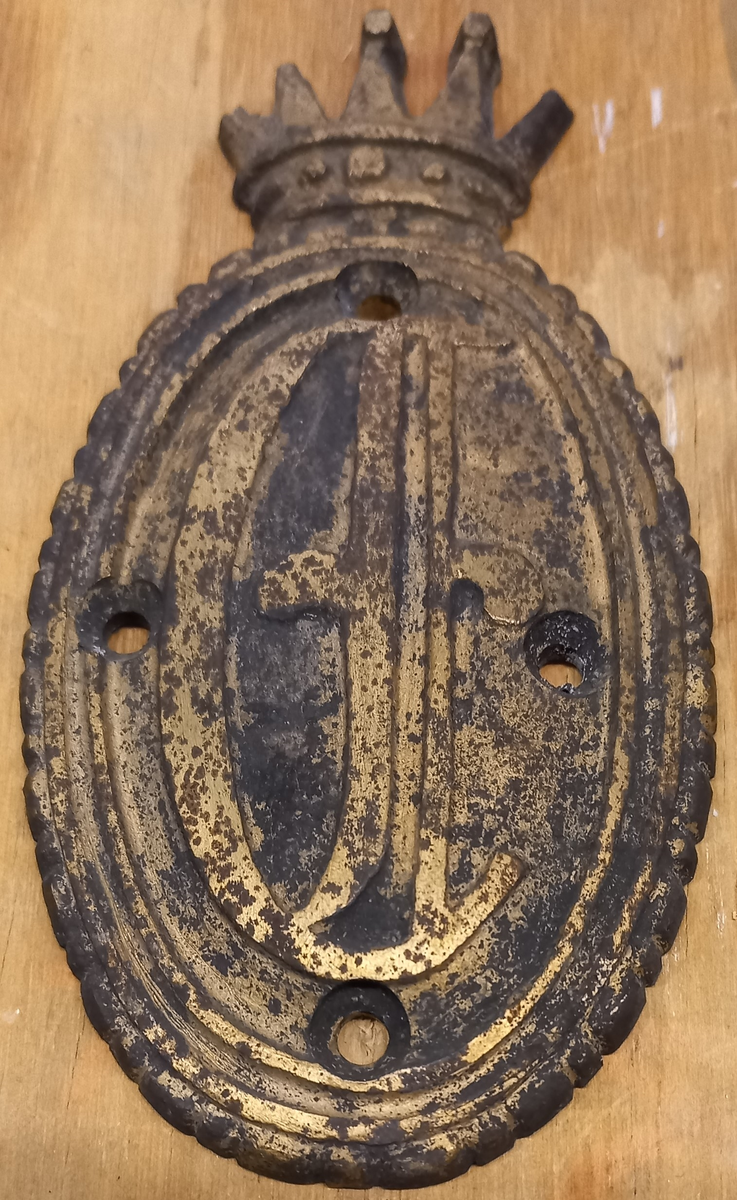 Stävmärke, krönt sköld, tillhört fregatten Eugenies båtar. Oval sköld med hertigkrona. I skölden ett E (tecknad figur). 4 stycken hål för skruvar. Förgylld.