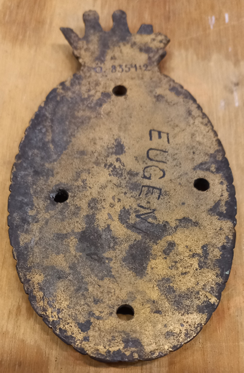Stävmärke, krönt sköld, tillhört fregatten Eugenies båtar. Oval sköld med hertigkrona. I skölden ett E (tecknad figur). 4 stycken hål för skruvar. Förgylld.