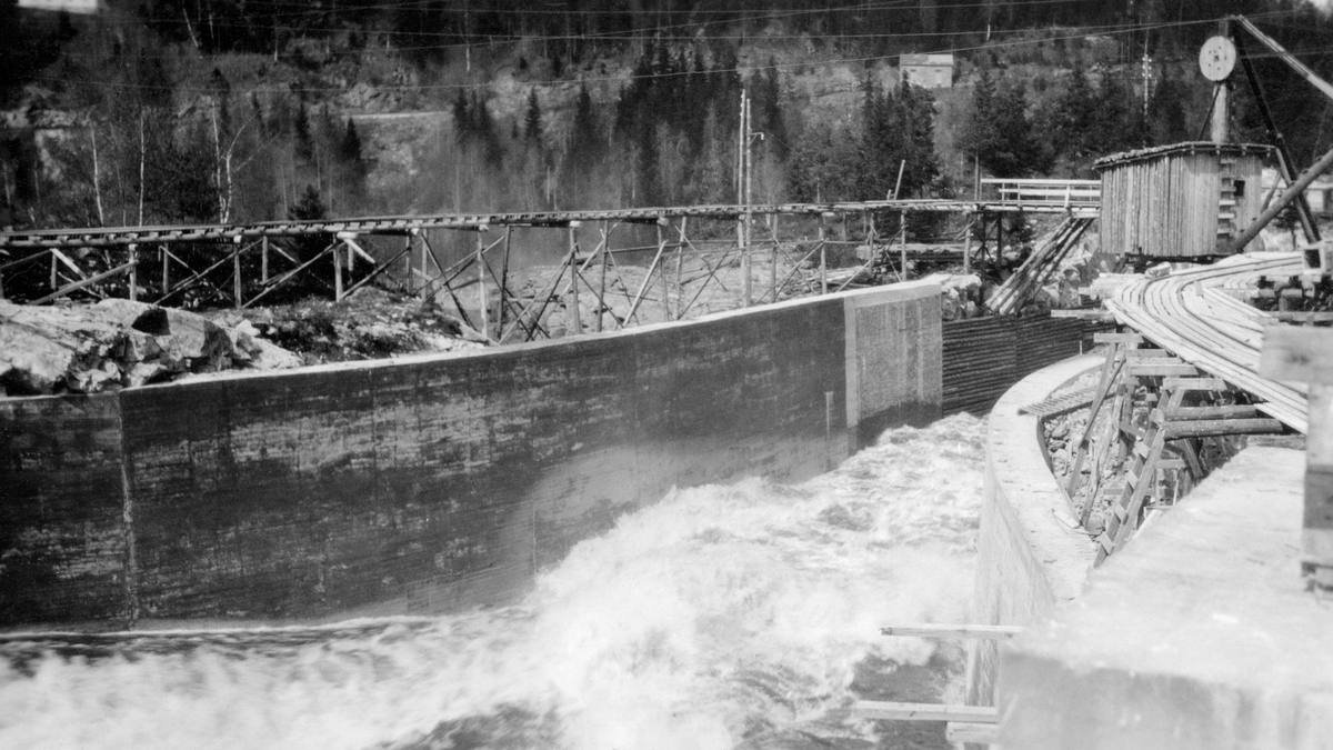 Fra tømmerkanalen ved Vamma i Askim i Østfold (nedre Glomma).  Denne kanalen ble innviet i 1927, etter at Fredrikstad Tømmerdirektion i elleve sesonger (1916-26) med vekslende hell hadde forsøkt å fløte de store tømmermengdene i denne delen av vassdraget gjennom ei underdimensjonert tømmerrenne litt lengre vest.  Dette fotografiet er antakelig tatt fra det 35 meter brede inntaket ned mot det videre kanalløpet, som var 12 meter bredt.  Derfor fant ingeniørene som planla anlegget det nødvendig å støpe høye betongvegger her.  Helt til høyre i bildet, bakenfor den buete betongskjermen mot kanalen ligger det ei kjørebru med skinnegang på et underlag av trebukker - antakelig en trallebane for å få fram materialer til byggearbeidene.  Ved siden av denne transportåren ser vi også et lite bordskur med pulttak.  På motsatt side av flomløpet og bak den buete betongskjermen der ser vi også en transportbane som kviler på trebukker, i dette tilfellet mye høyere over marka.  I bakgrunnen en delvis skogkledd bakkekam med en veg og et hus. 