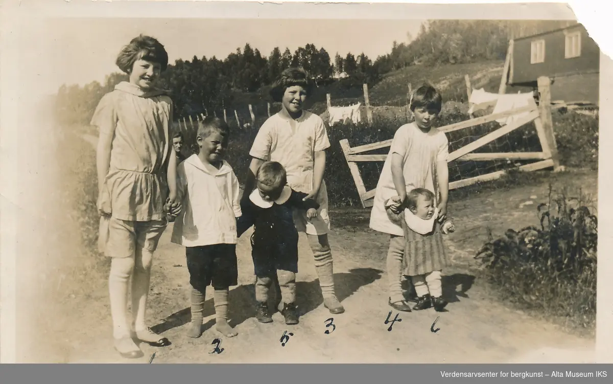 Seks barn står på en vei, de ser på kameraet og de eldre barna hjelper de yngste å stå, og bak dem ser vi hodet til en kvinne. Bak dem er det et hus, og det henger klær ute til tørk på gjerdet foran huset. Barna er nummeret, og navnene står på baksiden av bildet.