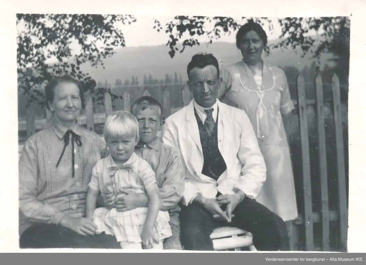 To voksne og to barn sitter på en benk, en kvinne står bak dem, de ser på kameraet og smiler. Bak dem er et gjerde og flere trær. 