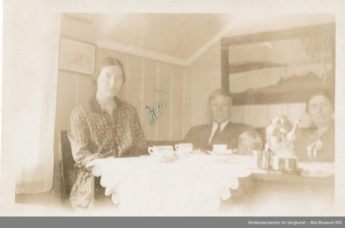 Tre voksne og et barn sitter rundt et bord. Bordet er dekket på med duk, tekopper og en kaffekanne. På veggen bak dem henger det et naturbilde. 