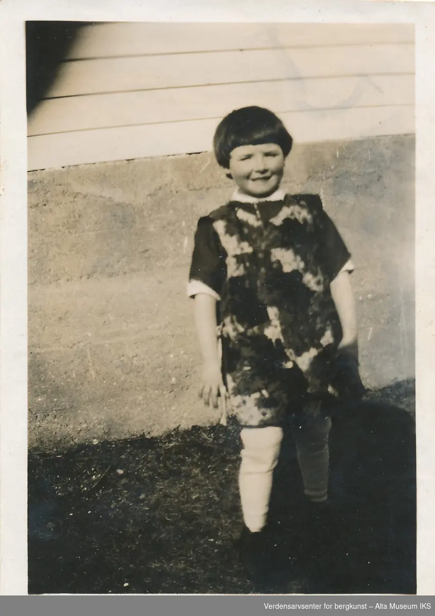 En jente står ute foran en husvegg og smiler til kameraet. Hun har på seg kjole og holder noe i hånden.