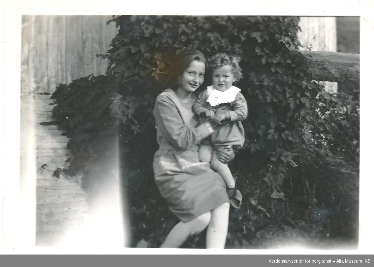En eldre jente holder en yngre gutt, de ser på kameraet, jenta smiler og gutten lager en grimase. De er ute foran en busk. På baksiden av fotografiet står det: "Berte og Lillegutt Gjrerdsen. Septbr. 1931"