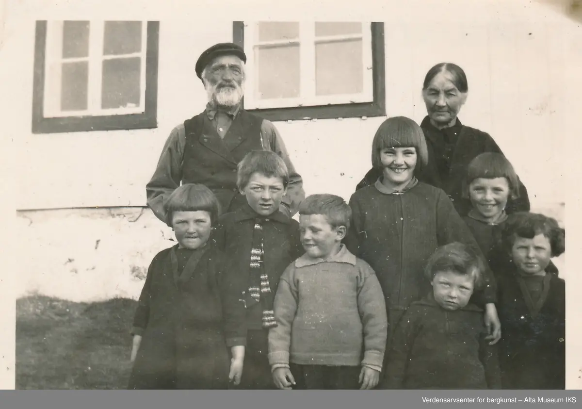 En gruppe mennesker står oppstilt ute foran en husvegg. En eldre mann og en eldre kvinne står bak en gruppe barn i ulike aldre. 