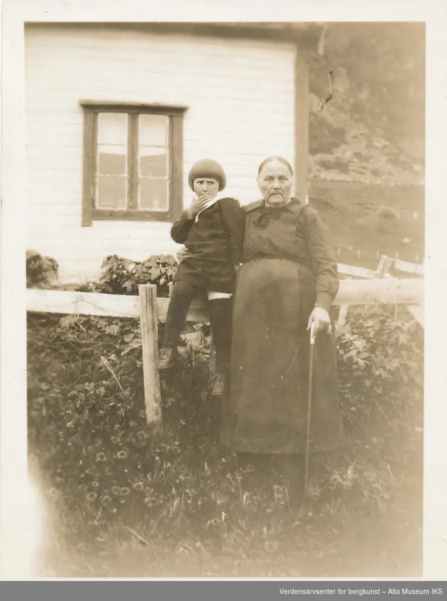 En eldre kvinne holder rundt et barn som sitter på gjerdet foran et hus. Kvinnen har på seg kjole og bruker stokk. På baksiden av bildet står en hilsen.