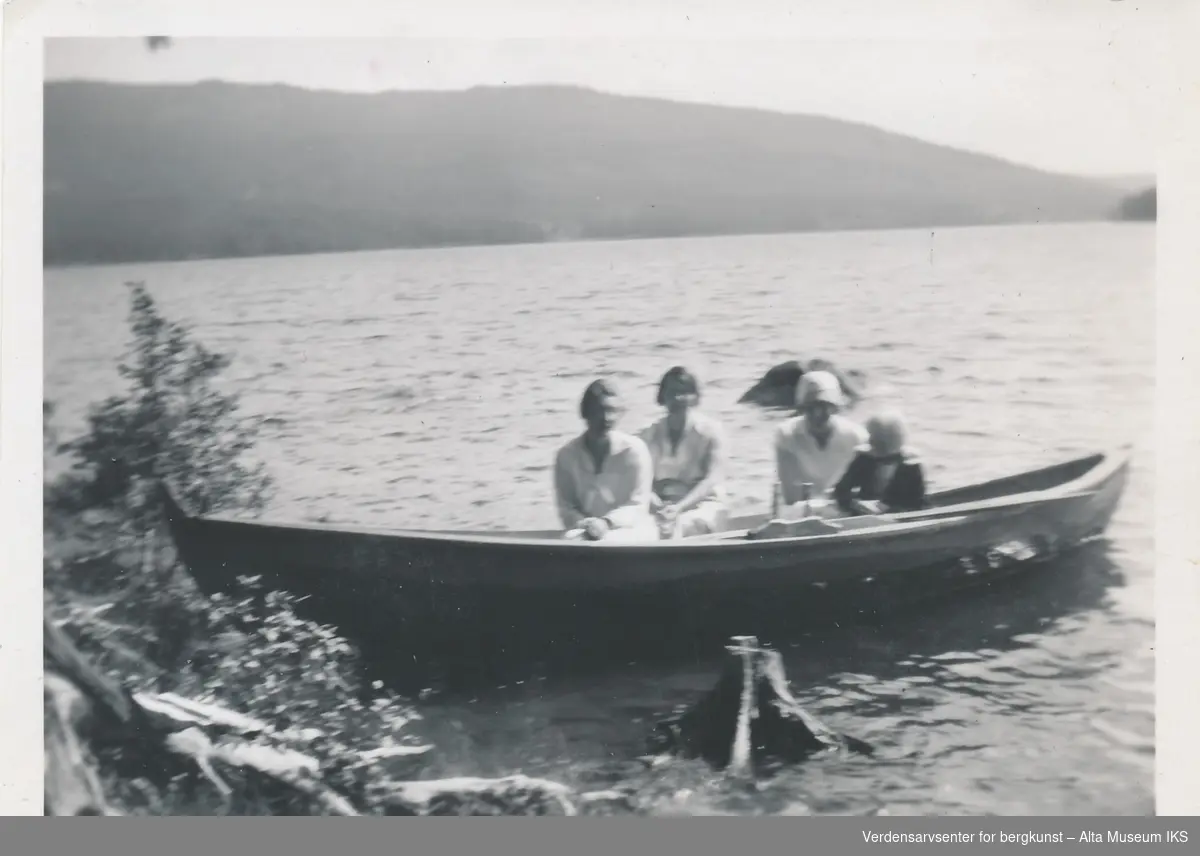 Tre kvinner og et barn sitter i en robåt i en innsjø, båten er nært land og noen busker. Bildet er tatt fra land, og kvinnene ser på kameraet. 