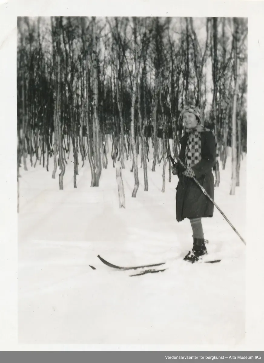 En kvinne står på ski i skogen. Hun har på seg skjerf og ser på kameraet. På baksiden av bildet står en liten hilsen.