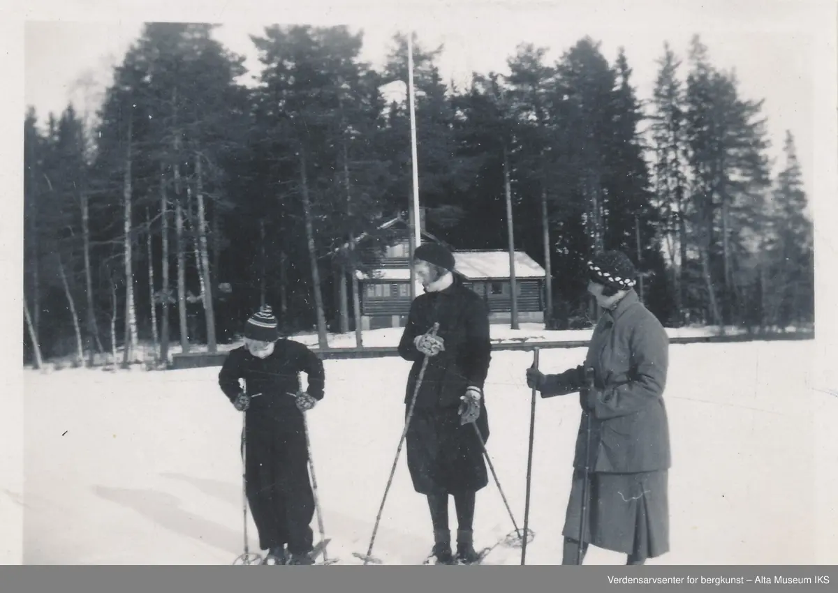 To kvinner og et barn står på ski på en slette foran en stor bygning i skogen. Framhytta.