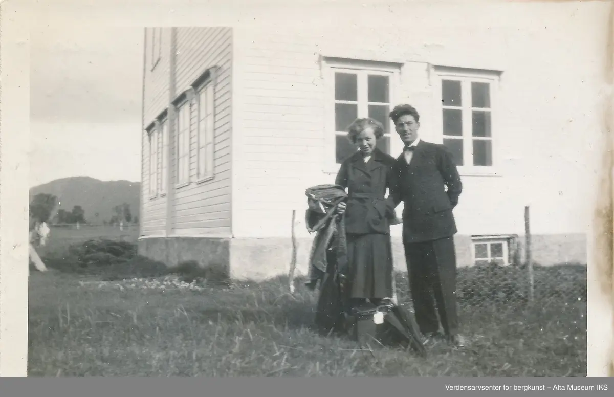 En mann og en kvinne står og holder rundt hverandre på gressplenen foran et hus. Bak dem er også en inngjerdet kjøkkenhage. På baksiden av bildet er det skrevet en hilsen. 