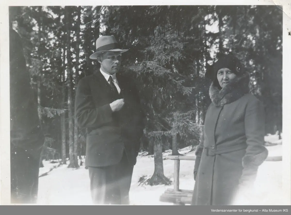 En mann med hatt og en kvinne i kåpe med pelskrage står ute i skogen, det er snø på bakken. Kvinnen ser på kameraet og mannen ser på henne. 