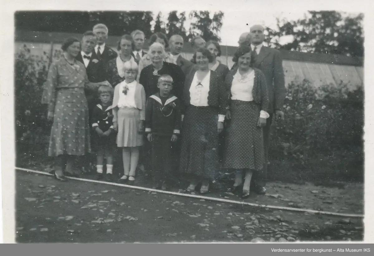 Gruppebilde, med kvinner, menn, barn, unge og eldre. De står stilt opp ute og de ser på kameraet og smiler. Det ligger blader på bakken, og bak dem er en busk og en bygning. 