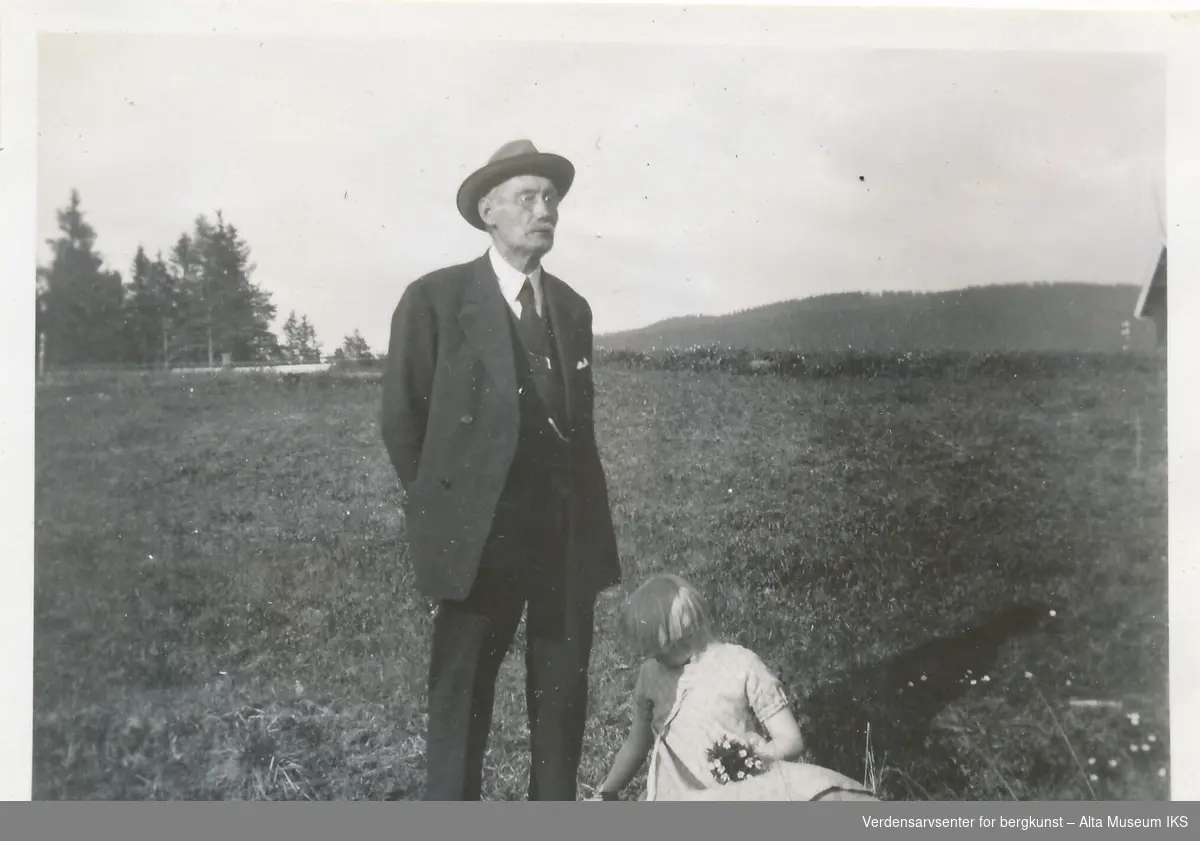 En eldre mann med hatt og briller står i en eng, han har hendene foldet bak seg og ser forbi kameraet.  Ved beina hans sitter et barn i kjole og plukker blomster, hun holder den lille buketten hun jobber på i fanget. Bak dem strekker engen seg til trærne, og skogen tar over. 