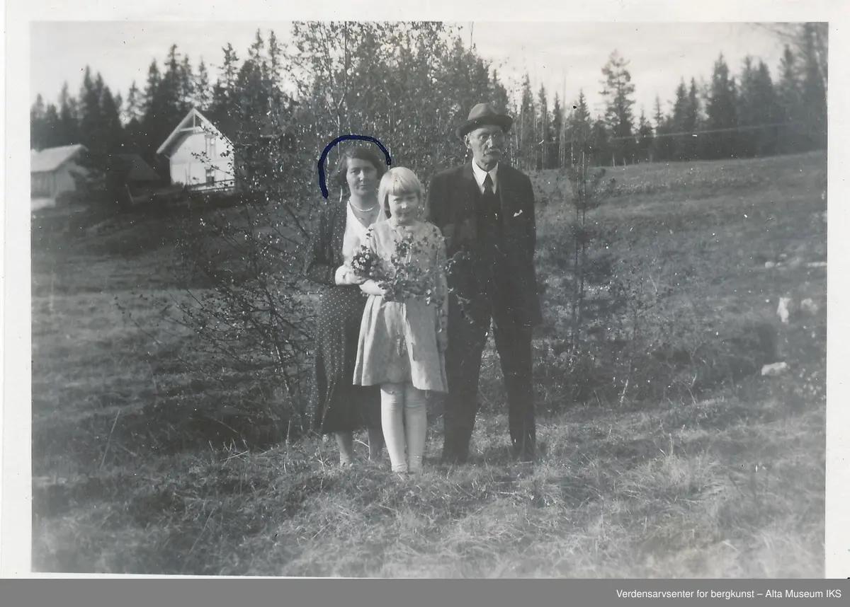En eldre mann med hatt og briller, en kvinne i kjole og et barn som holder en blomsterbukett står stilt opp sammen på en eng foran en busk. De ser på kameraet og smiler. Bak dem strekker engen seg til toppen av bakken før skogen tar over, og øverst i bakken står flere bygninger. Det er tegnet på en halvsirkel over kvinnens hode, og et minus tegn i høyre hjørnet. På baksiden av bildet står også en beskjed.