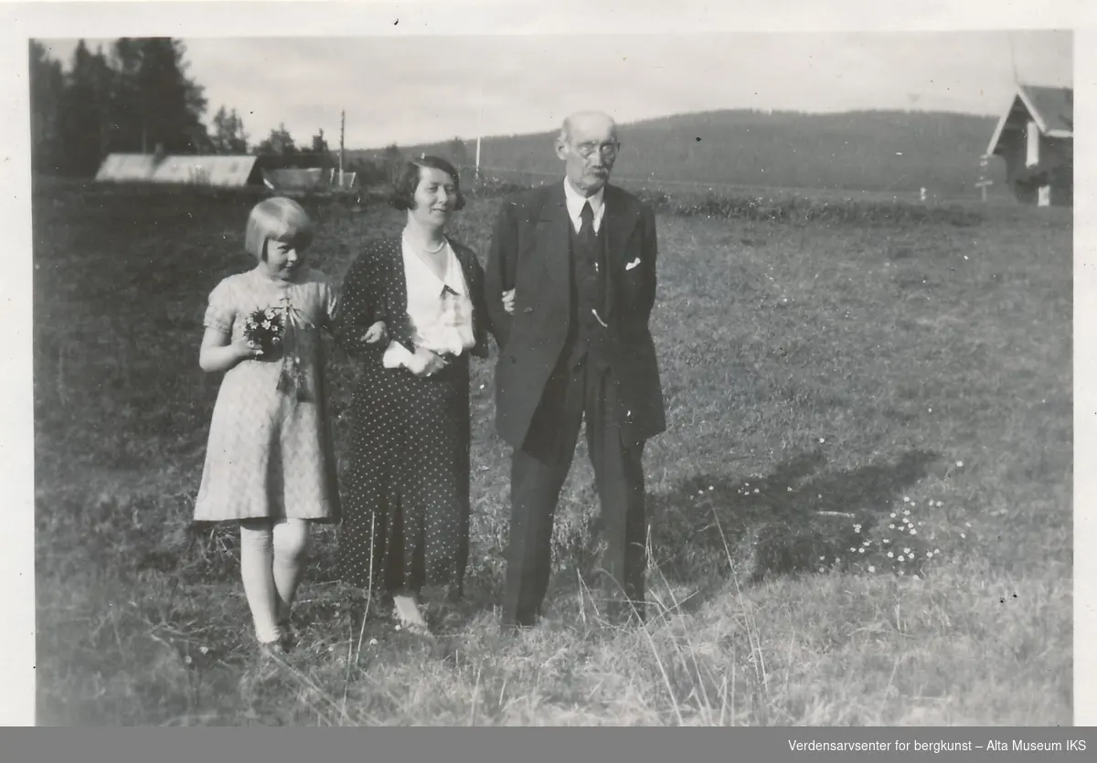 En eldre mann med hatt og briller, en kvinne og et barn som holder en blomsterbukett står stilt opp sammen på en eng, de holder rundt hverandre. Bak dem øverst i bakken står en bygning, og bak et gjerde starter skogen. 