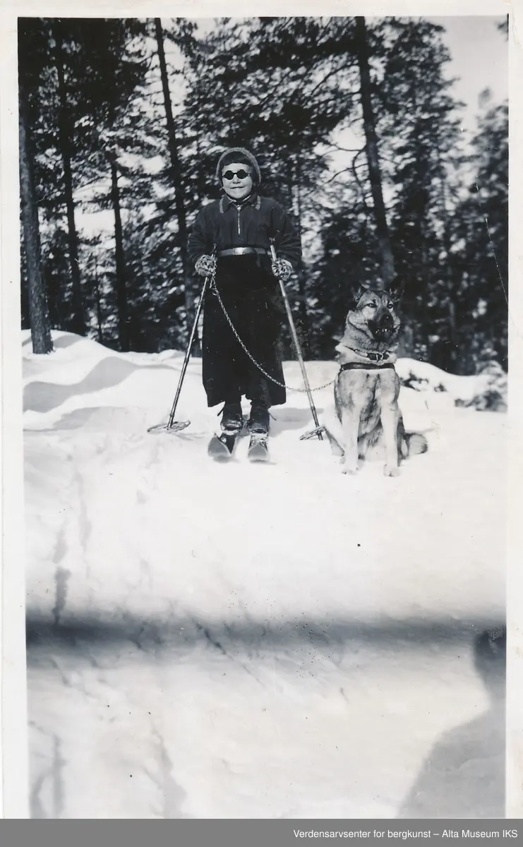En jente står på toppen av en bakke på ski, hun holder en hund som sitter ved siden av henne i bånd og hun smiler stort til kameraet. På baksiden av bildet er det skrevet en gratulasjon. 