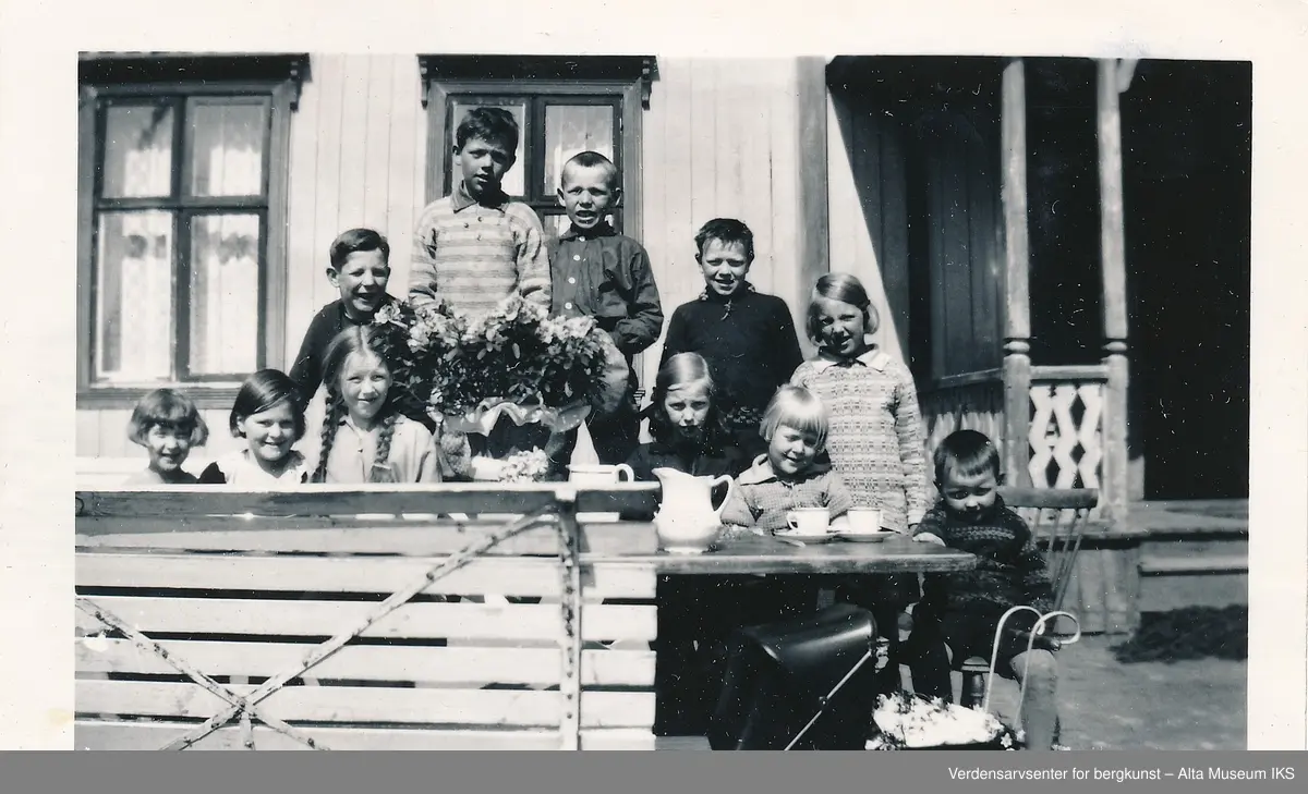 En gruppe barn sitter ute rundt et hagebord dekket på med kaffekopper og blomster. Ved siden av bordet står en liten dukkevogn, og bak dem er et hus. Alle barna ser på kameraet og smiler. 