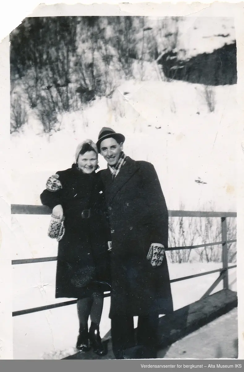 En mann og kvinne holde rundt hverandre og lener seg mot rekkverket til en bro, de smiler til kameraet. Det er snø på bakken rundet dem, og de har på seg votter og frakk og kåpe. 