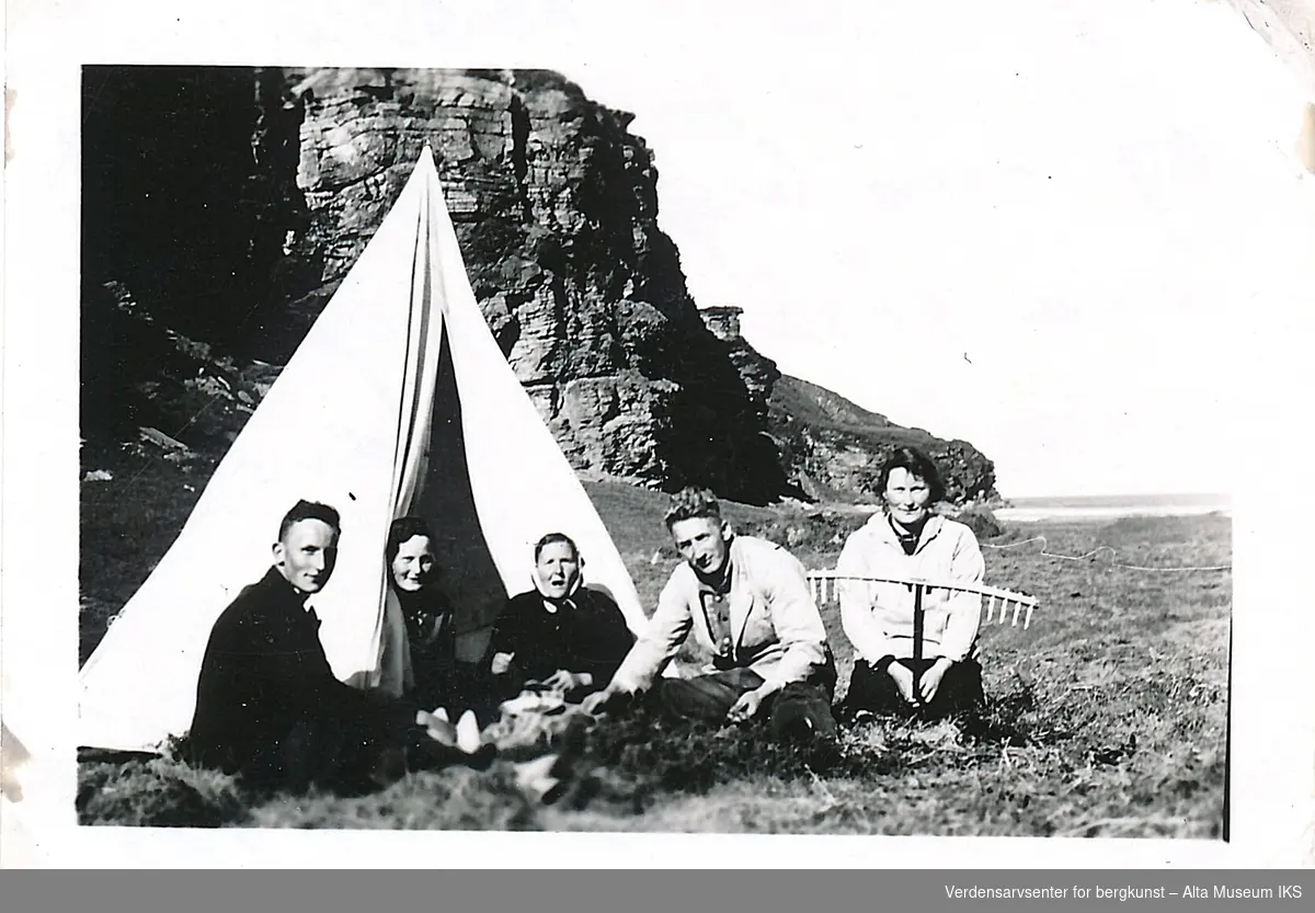 En gruppe mennesker sitter rundt et hvitt telt på en slette foran en fjellside. Den ene kvinnen holder en rive foran seg. På baksiden av bildet er det skrevet en hilsen.