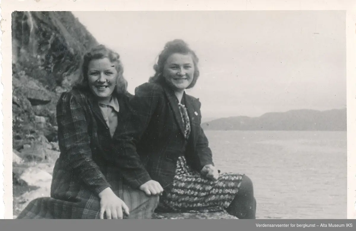 To kvinner sitter sammen på en stein i fjæren foran havet ut mot fjorden. De lener seg på hverandre og smiler til kameraet. 