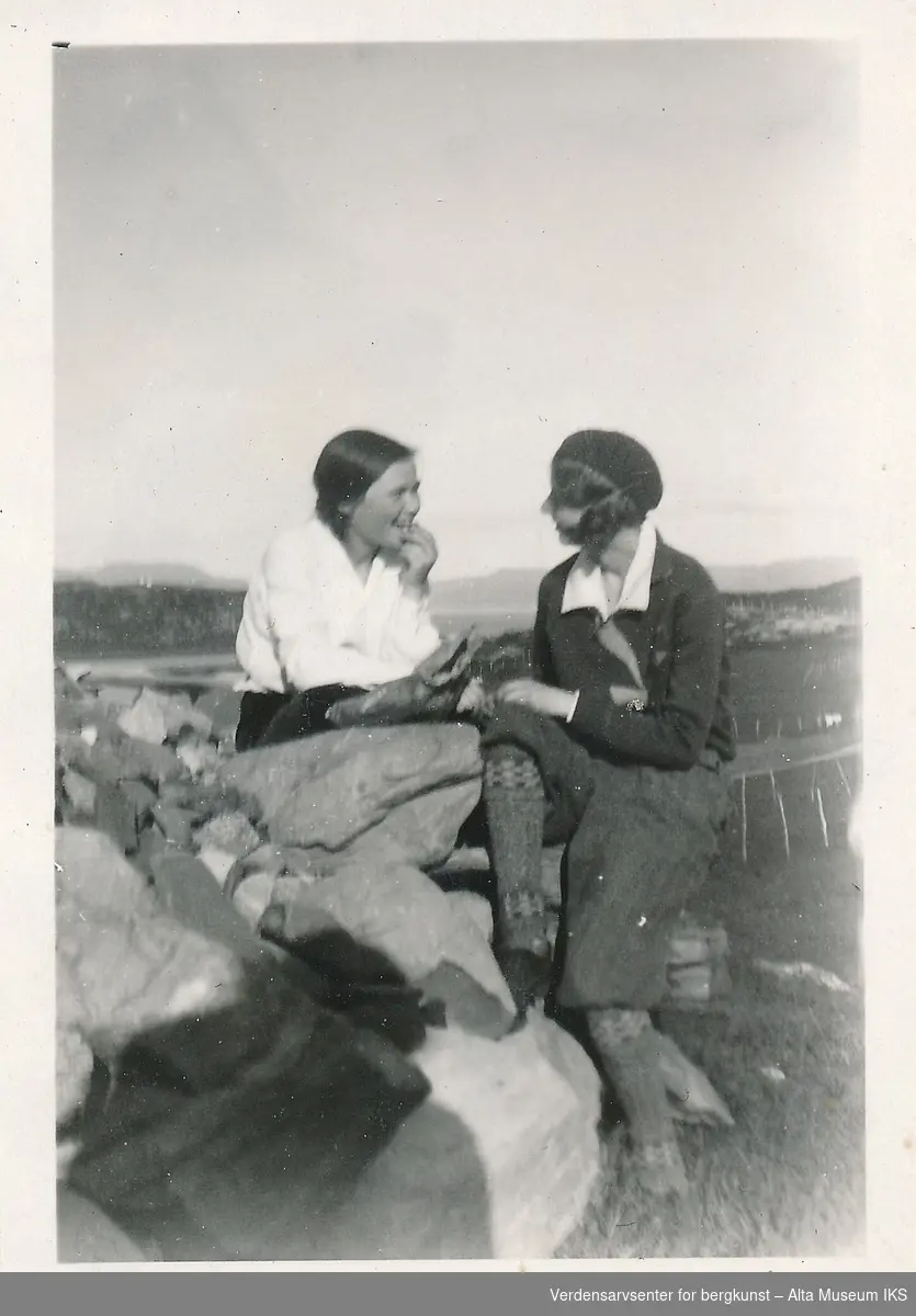 To kvinner sitter på et steingjerde, og de ser på hverandre. Bak dem er det utsikt over et gjorde og fjorden. 