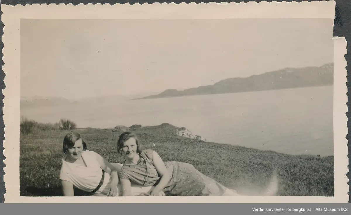 To kvinner ligger i gress landskap, de lener seg på hverandre og smiler til kameraet. Bak dem har de utsikt over havet og fjorden. 