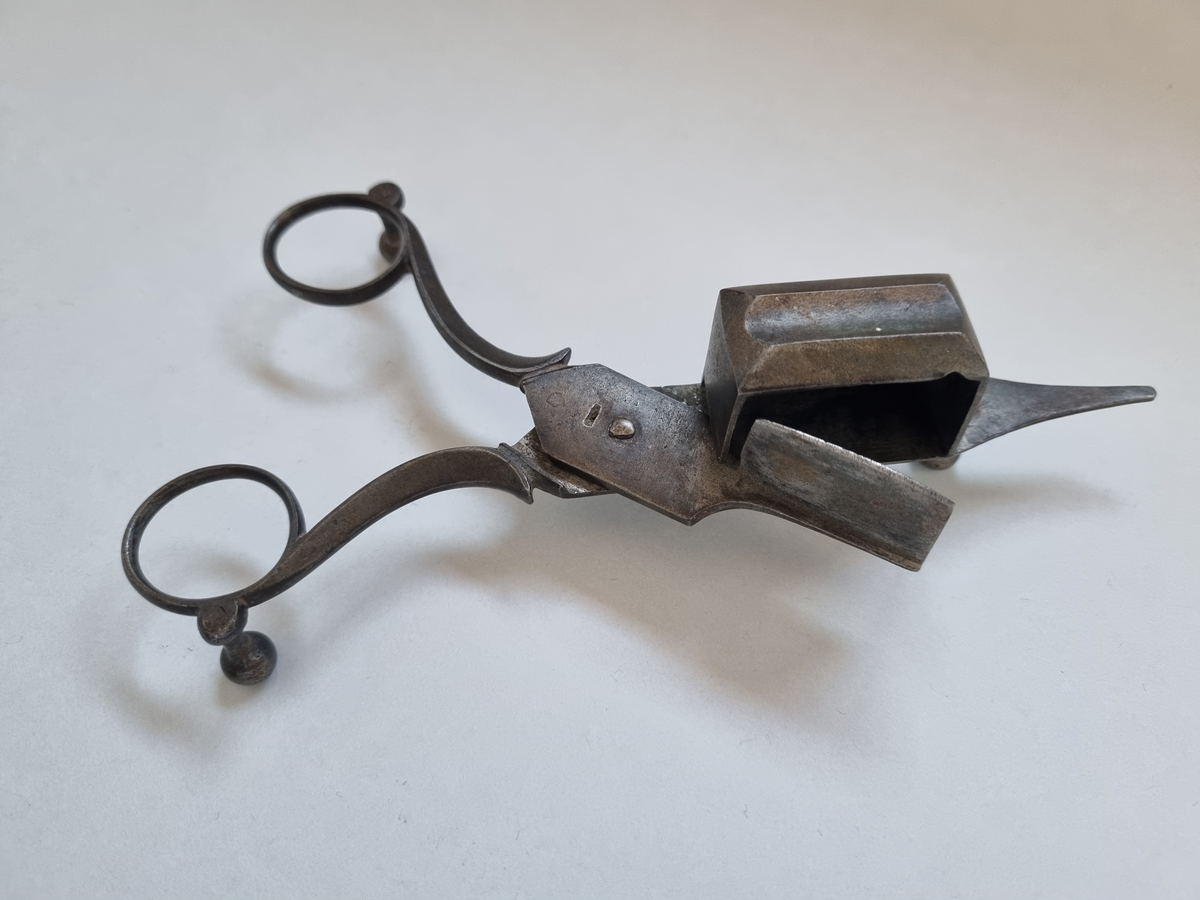 Ljussax i brons. Ljussaxen användes innan den självförintande veken uppfanns. Behållaren är avsedd för den avklippta veken. Spetsen är till för att räta upp veken med. Saxen vilar på tre fötter för att inte sota ner underlaget.
