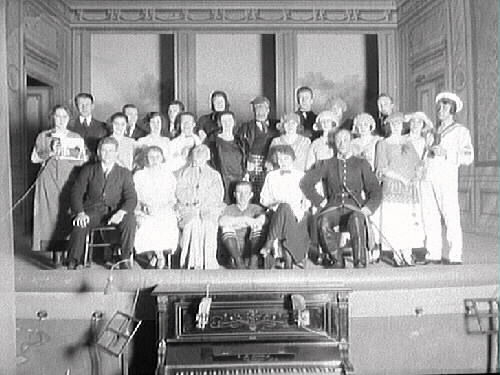 Ensemblen i nyårsrevyn "Än leve de gamla gudar!" 1924 på Varbergs teater. Efter premiären den 1 januari fick revyn först ett ljumt mottagande i recensionen av "Brutus" i Varbergsposten: "Anrättningen befanns vara skäligen mager...". Men efter viss omarbetning och mer repetitioner till föreställningen en vecka senare fick den idel lovord. Bland artisterna fanns kuplettsångarna Ernst Dahlqvist och herr Rosengren. Komikern B G Möllerström fick enligt Brutus publiken att tjuta av förtjustning åt roller som "lantliga Truls", slaktardräng, beväring och kapellmästare. Den verklige kapellmästaren hette Dahlgren och fick beröm, liksom "Jassflickorna" och "Valkyriorna". Men allra mest imponerade M Aschjem som både tränat in baletterna och målat dekorerna: "Aldrig förut har en lokalrevy här kunnat ståta med så pampiga dekorationer och finfina dräkter." Här ses bl a en sjöman, en fotbollsspelare, en kvinna hållande en vargfigur på hjul i snöre och självaste Karl XII. Lokalrevyn gjordes till förmån för Varbergs Gymnastik- och Idrottsförening.
Bild 1: Tonat foto (F9358) med Björkströms logotyp monterat i omslag.
Bild 2: Glasplåt, nedanför scenen ses piano och notställ. 
(Se även bildnr GB2_2778-2785)