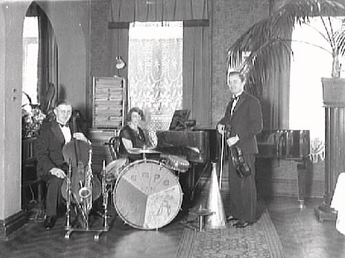 Lisa Lampas trio, bestående av två män och en kvinna, som sitter vid flygeln. Mannen till höger med fiol är Fritz Moreau och till vänster sitter en man med cello. Mellan herrarna står även trummor och blåsinstrument. Musikerna är fotograferade i matsalen på Varbergs Hotell. (Se även bildnr GB2_4013)