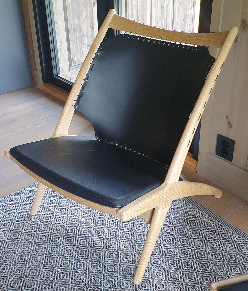 Lenestol av tre med svart skinn som er strukket som et segl i ryggstøtten. Svart sittepute av skinn. Stolbeina og ryggstøtten er formet som et kryss.