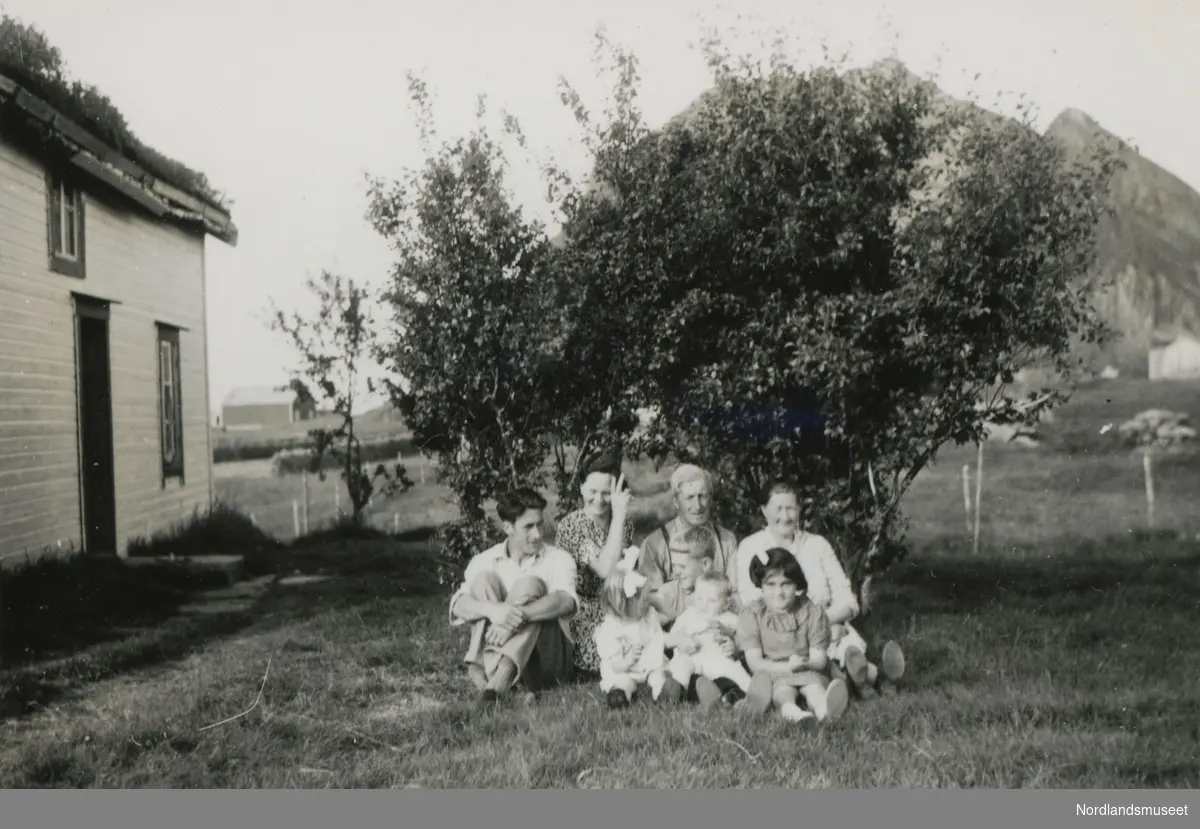 Familie som sitter på bakken utenfor huset sitt i Bø på Engeløya. De voksne fra venstre: Karl Meyer Kristensen (f. 1920) og kona Alma (f. 1915). Videre er Karls foreldre Magnus Alfred (f. 1892) og Ingeborg (f. 1895). Ungene er Marta og Edgar Kristensen, Trygve Sandvik og Annhild Slydal.  Alma, pikenavn Iversen, kom fra Lund i Leiranger.