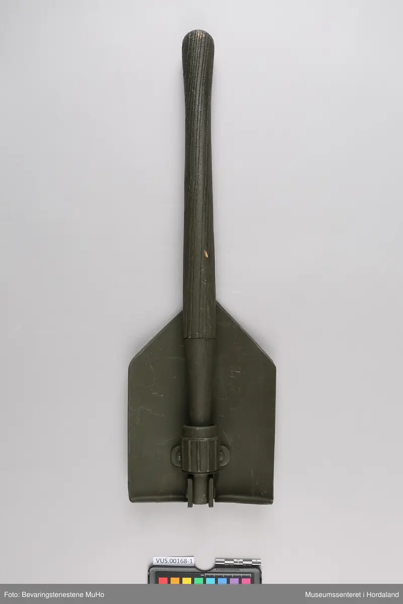 Militærgrønn feltspade og spadeveske. Feltspaden er av metall med skaft av tre. Spadebladet er brettet inn bak skaftet og har en skruelås. Spadevesken er av bomullslerret. Den består av en oval bakdel med påsydd lomme og metallknapp. Lommen har hull nederst som spadeskaftet kan stikke ut av.