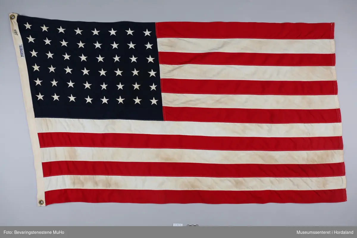 Stort, rektangulært flagg med tretten røde og hvite striper, samt 48 stjerner på blå bakgrunn i øvre venstre hjørne. Tilhørende flagget er en todelt flaggstang av tre og en massiv flaggfot av metall.