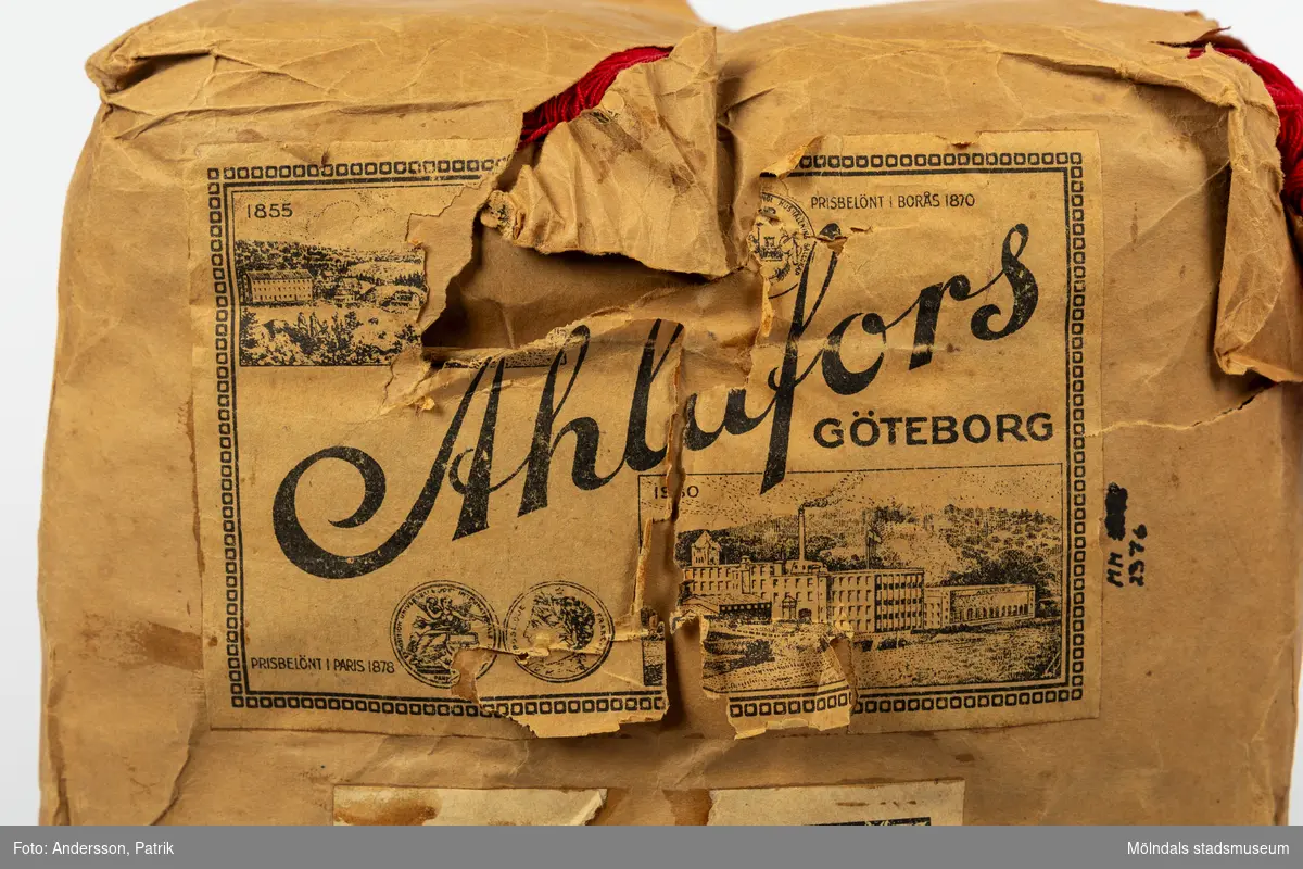 En förpackning med rött bomullsgarn (äkta rödfärg nr 39). Garnmängden (2 kg?) paketerat i brunt omslagspapper, med snöre runt om. Tre olika etiketter från Ahlafors är påklistrade. Pappret är trasigt.Tidigare sakord: garn i förpackning.