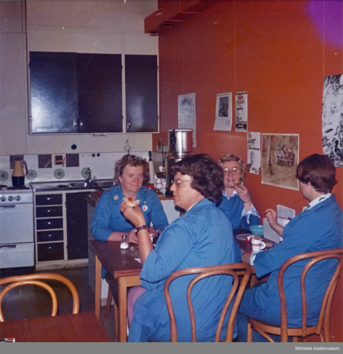 Ett kök/fikarum, Domus vid Nya torget senast 1976. Fyra kvinnor, iklädda blå rockar, sitter vid ett bord och fikar. På den röda/orangefärgade väggen bredvid kvinnorna sitter olika bilder uppsatta. I bakgrunden ses kökets brunmålade luckor och lådor.