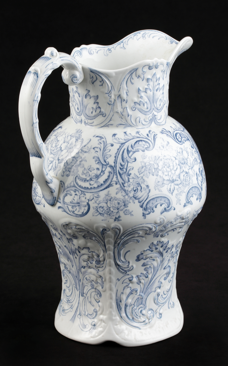En handkanna av vitt flintporslin. Balustermodell med S-format handtag.
Dekor av blommor och akantusblad med blå kanter, i relief.
Ingår i tvättservis.
