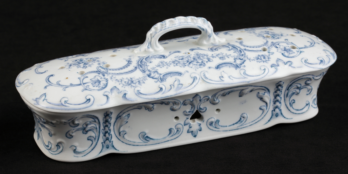 En avlång kamask med lock, av vitt flintporslin. 
Tre mindre fack invändigt. På sidorna tre mindre hål, varav ett hjärtformat. Lufthål (2x7) i locket.
Dekor av blommor och akantusblad med blå kanter, i relief. 
Ingår i tvättservis.