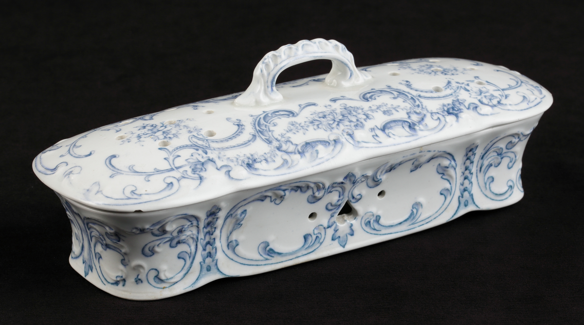 En avlång kamask med lock, av vitt flintporslin. 
Tre mindre fack invändigt. På sidorna tre mindre hål, varav ett hjärtformat. Lufthål (2x7) i locket.
Dekor av blommor och akantusblad med blå kanter, i relief. 
Ingår i tvättservis.
