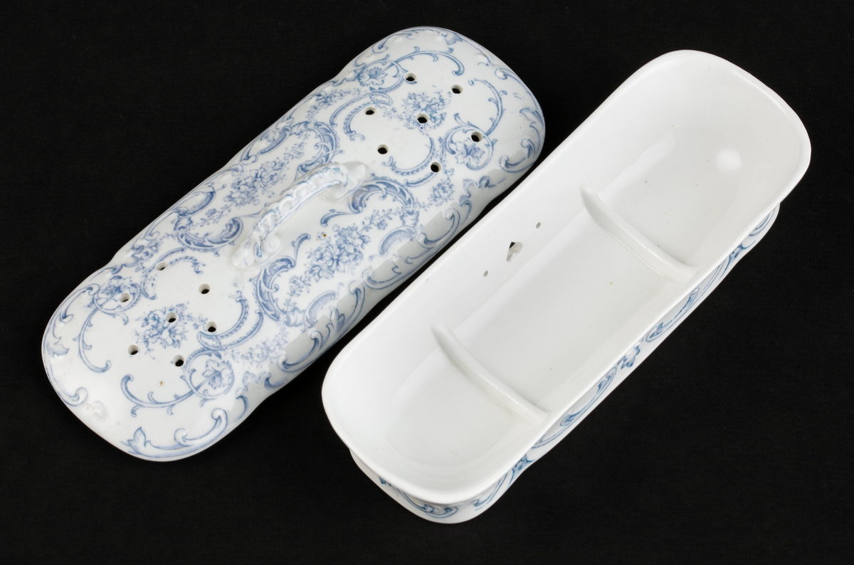 En avlång kamask med lock, av vitt flintporslin. 
Tre mindre fack invändigt. På sidorna tre mindre hål, varav ett hjärtformat. Lufthål (2x7) i locket.
Dekor av blommor och akantusblad med blå kanter, i relief. 
Ingår i tvättservis.