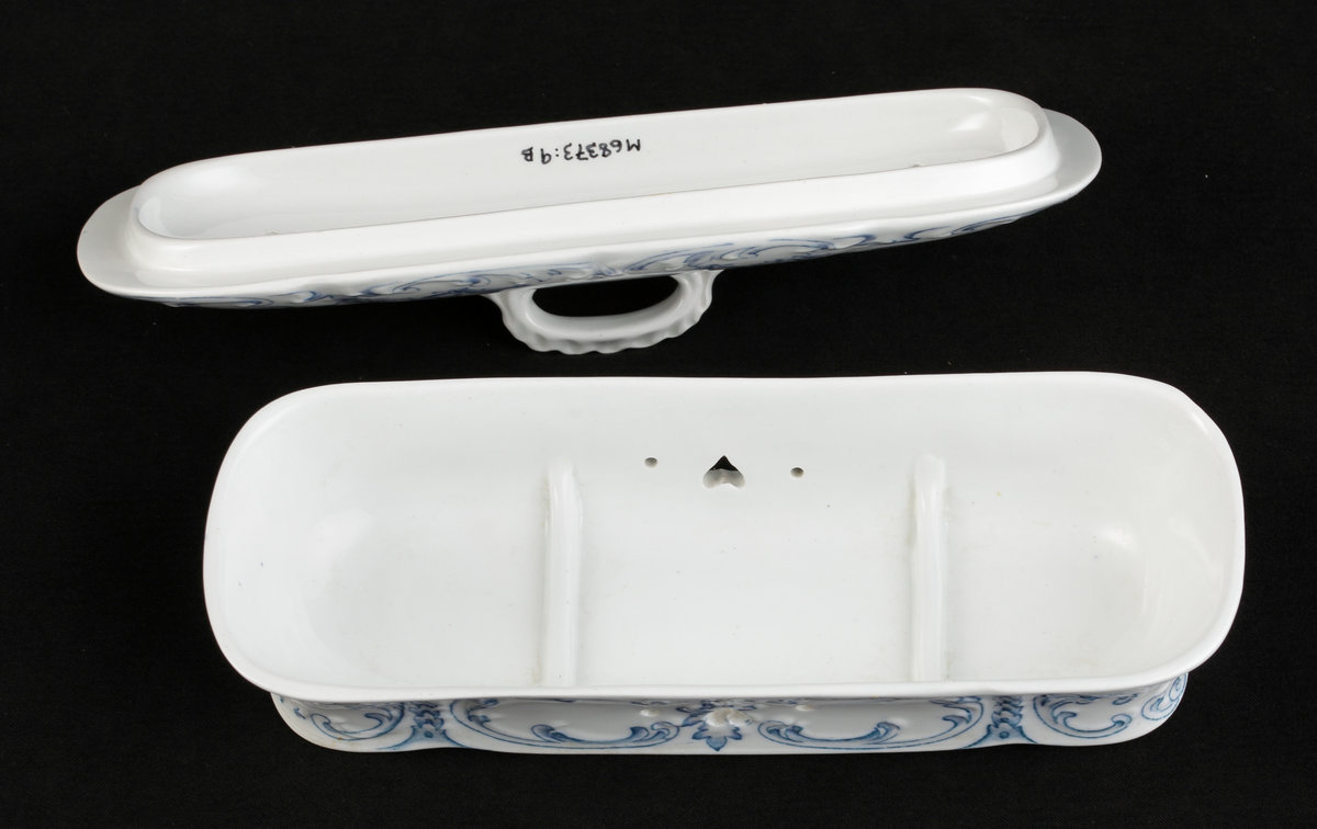 En avlång kamask med lock, av vitt flintporslin. 
Tre mindre fack invändigt. På sidorna tre mindre hål, varav ett hjärtformat. Lufthål (2x7) i locket.
Dekor av blommor och akantusblad med blå kanter, i relief. 
Ingår i tvättservis.
