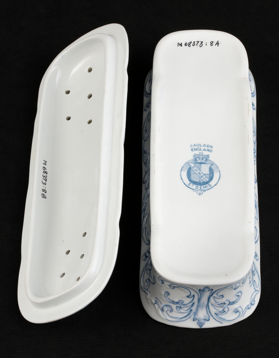 En avlång kamask med lock, av vitt flintporslin. 
Tre mindre fack invändigt. På sidorna tre mindre hål, varav ett hjärtformat. Lufthål (2x7) i locket.
Dekor av blommor och akantusblad med blå kanter, i relief. 
Ingår i tvättservis.