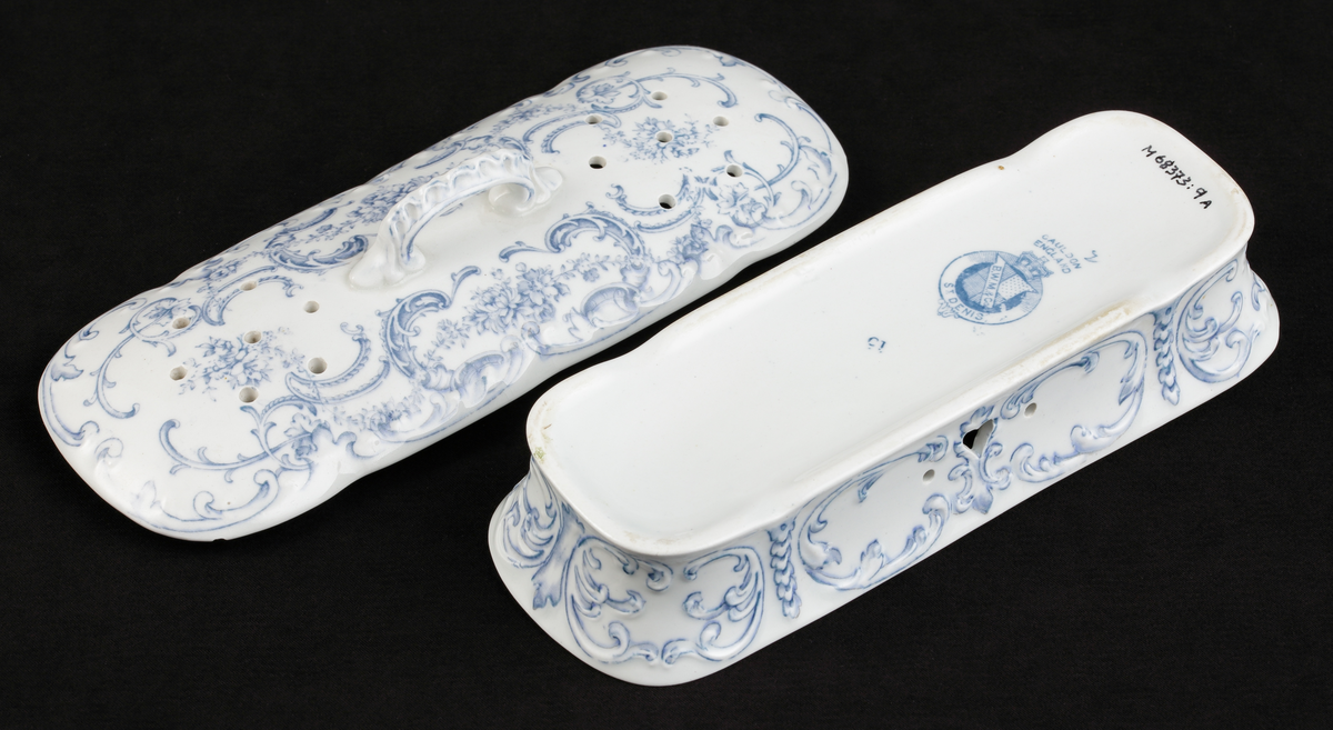 En avlång kamask med lock, av vitt flintporslin. 
Tre mindre fack invändigt. På sidorna tre mindre hål, varav ett hjärtformat. Lufthål (2x7) i locket.
Dekor av blommor och akantusblad med blå kanter, i relief. 
Ingår i tvättservis.
