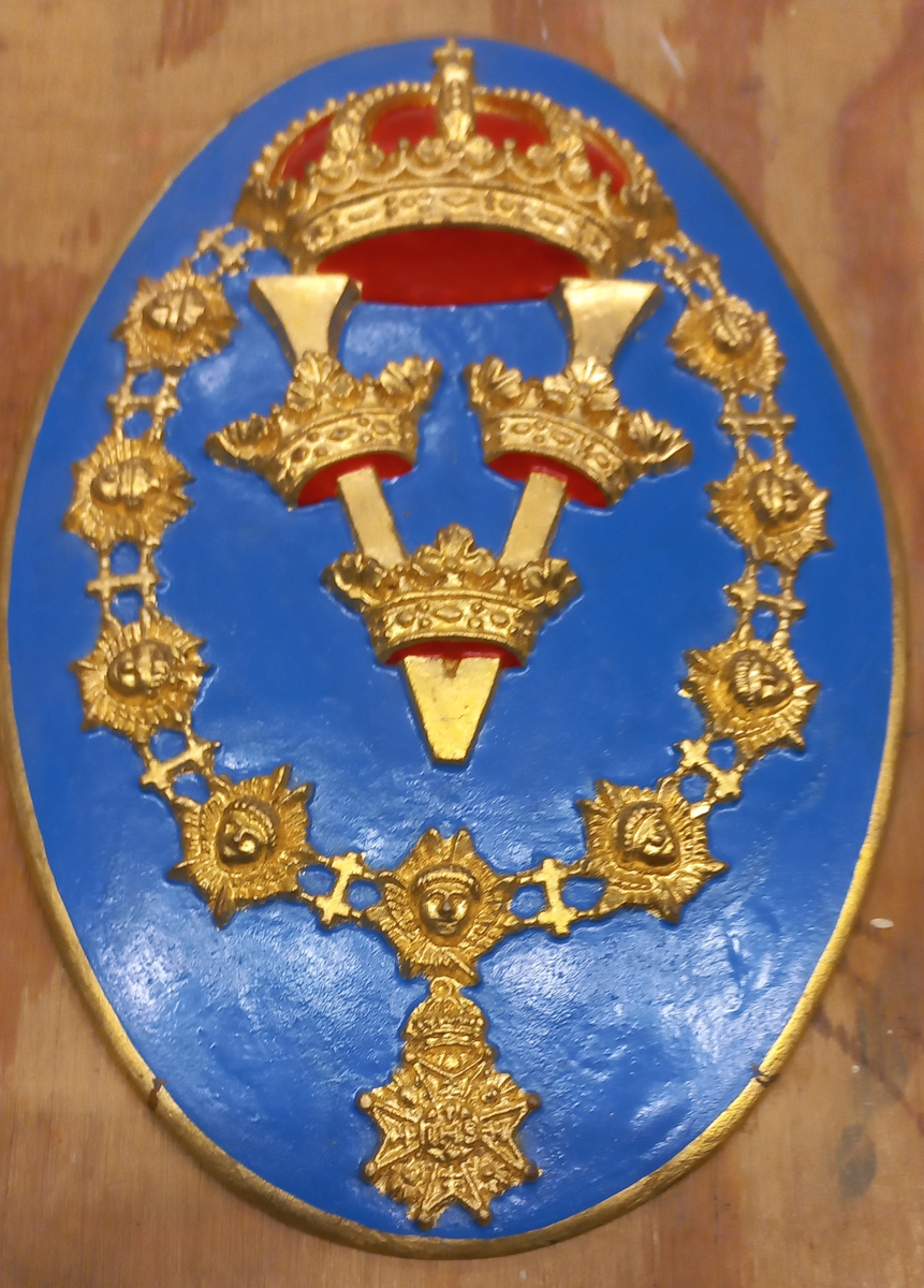 Oval blå sköld med Gustaf V:s namnskiffer inom serafimerkedja och krönt med Kunglig krona. H.M. Pansarskepp Gustav V:s vapen