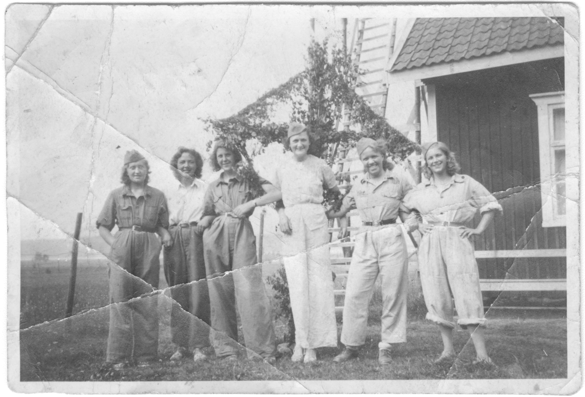 Gruppfotografi av sex luftbevakningslottor framför midsommarstång vid hus, midsommaren 1941.
Under luftbevakning i Rud, Värmland.