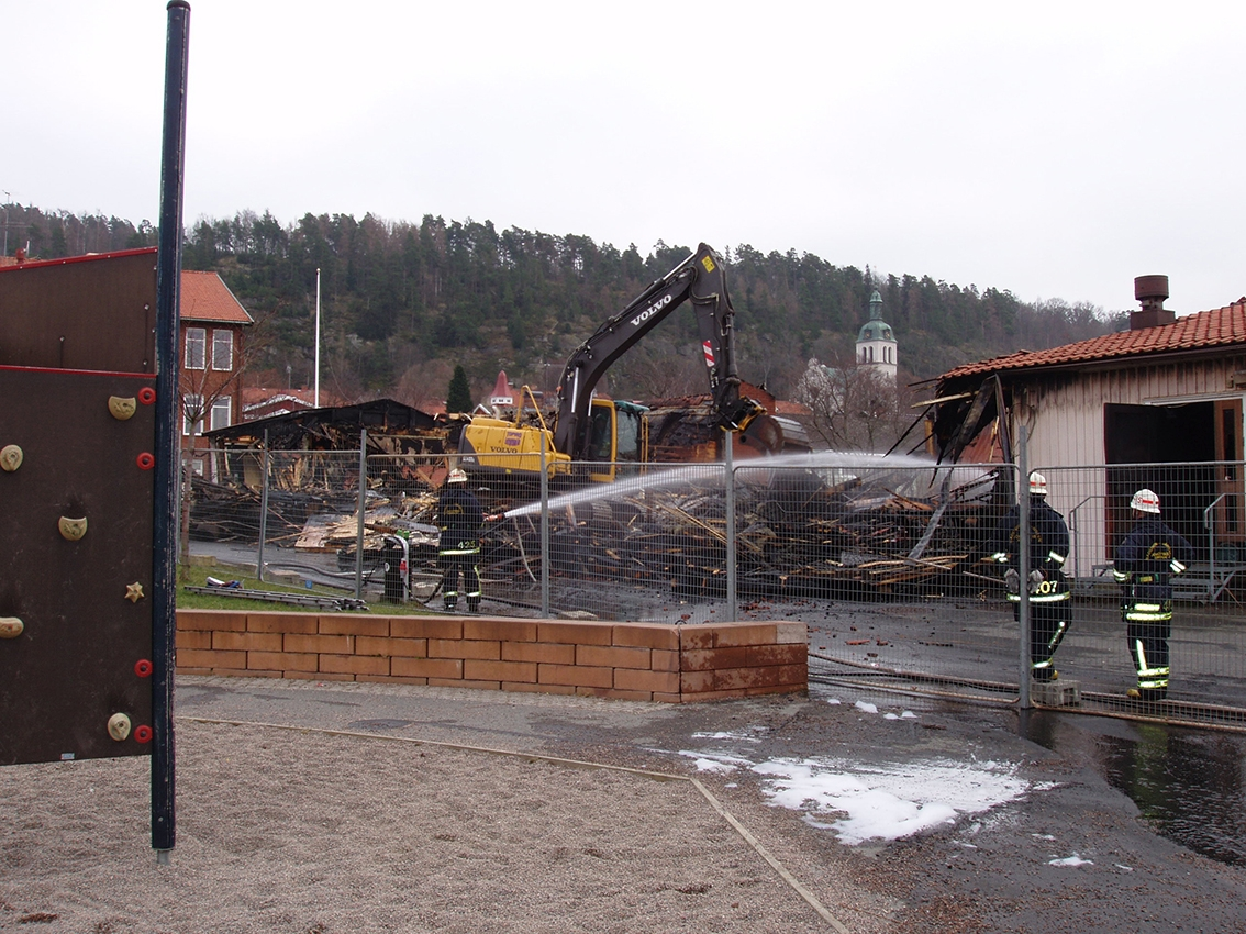 En grävmaskin river i en brandskadad skolbyggnad och en brandman sprutar vatten på arbetsområdet.